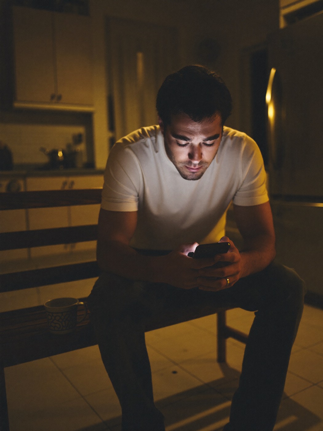 A father sits in a dark kitchen late at night, face lit by his phone